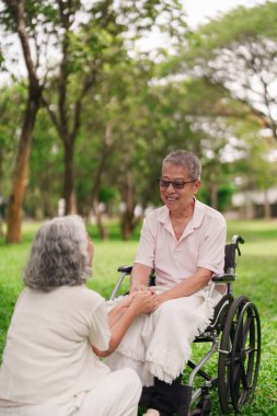 Asyalı yaşlı çift, özel an parkını paylaşıyor. Kadın, tekerlekli sandalyeye oturan adamı rahatlatıyor. Sevecen el ele tutuşmalar sevgi desteğini temsil eder. Birlikte emeklilik yılları.