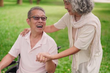 Parktaki yaşlı, mutlu Asyalı çift. Tekerlekli sandalyede partnerin desteğiyle oturuyor.