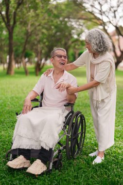 Sevgili Asyalı eş, yaşlı kocasına dışarıda tekerlekli sandalyede yardım ediyor. Emeklilik parkında bakım desteğini gösteriyor.