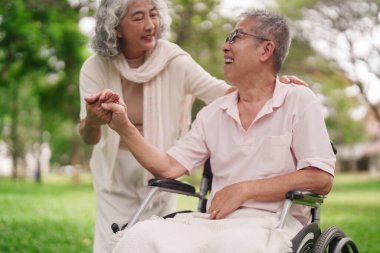 Kıdemli Asyalı kadın, mutlu bir ortağa yardım ediyor. Tekerlekli sandalyesini açık havada kullanıyor. Şefkat ve engelli bakımı gösteriyor.