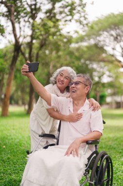 Asyalı yaşlı çift, dışarıda mutlu anları yakalıyorlar. Tekerlekli sandalyede oturan yaşlı adam karısı arkada durmuş, fotoğraf çekiyor.