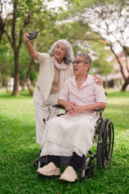 Asyalı son sınıf çift akıllı telefon özçekimini park ediyor. Tekerlekli sandalyedeki kadın eğleniyor. Mutlu emeklilik yaşam tarzı sağlık desteği aşkı.