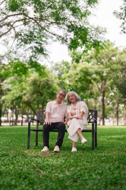 Neşeli yaşlı Asyalı çift parktaki bankta dinleniyor. Mutlu emeklilik hayatı sahneleri güzel yeşil açık havada barışçıl bağlar kurarak.