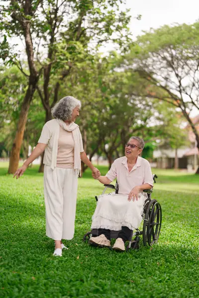 'Asyalı yaşlı bir çift parkta birlikte yürümekten zevk alıyor. Kadın tekerlekli sandalyedeki adama yardım ediyor. Mutlu emeklilik, yaşam destek, ilişki yaşlanma yolculuğu 