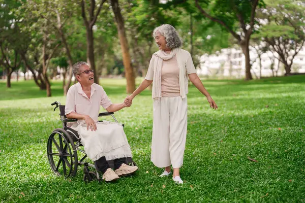 Asyalı son sınıf çifti mutlu bir gülümseme ile parkın tadını çıkaran kadın tekerlekli sandalyedeki adama yardım ediyor.