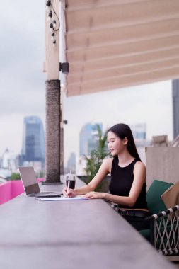 Güçlü Asyalı kadın şehrin çatı katındaki kafede kendine güvenerek çalışıyor. Modern bir şehir görüşünde dizüstü bilgisayarını kullanarak önemli notlar yazıyor..