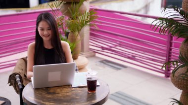 Kendine güvenen Asyalı kadın canlı bir kafede uzaktan çalışmaktan hoşlanıyor. Dizüstü bilgisayarına odaklanmış bağımsız bir ruhla başarı yaratıyor..