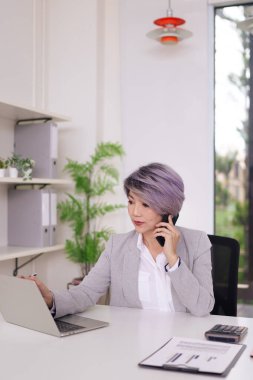 Profesyonel Asyalı kadın, bilgisayarını kullanarak ve akıllı telefonuyla çağdaş ofis ortamında iş yükünü aktif olarak yönetiyor.