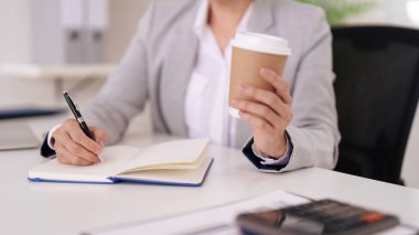 Yetişkin bir profesyonel, organize bir ofis ortamında detaylı notlar alıyor. Sıcak bir içecekle bir sonraki adımları planlama ve hazırlama görevlerine odaklan.