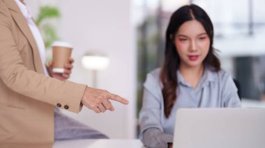 Profesyonel Asyalı kadınlar iş birliği yapıyor. Yetişkin bir iş kadını bilgisayarda çalışan bir meslektaşını işaret ediyor. İş başarısı için modern bir kurumsal ortamda takım çalışması rehberliği