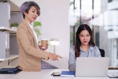 Deneyimli Asyalı yönetici genç meslektaşına dizüstü bilgisayarda rehberlik ederek parlak kurumsal ofis ortamında profesyonel gelişim ekip çalışmasını ve başarısını teşvik ediyor.