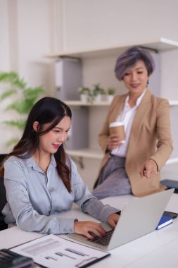 İki Asyalı profesyonel kadın verimli bir ofis işbirliği içinde. Tecrübeli bir akıl hocası, genç bir meslektaşına, modern bir iş yerinde dizüstü bilgisayardaki verileri gözden geçirmesinde yardımcı olur..