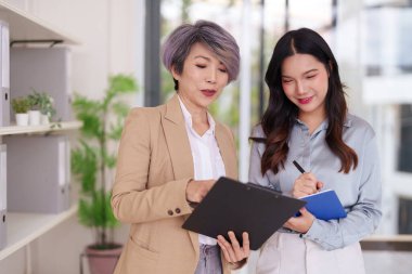 İki Asyalı iş kadını parlak, modern bir ofiste iş birliği yapıyorlar. Deneyimli bir yetişkin akıl hocası genç bir profesyonel değerlendirme planına rehberlik eder..