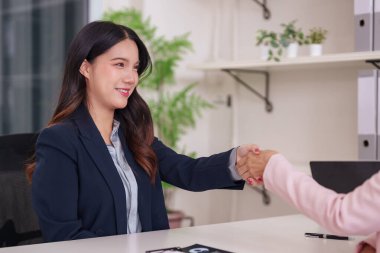 Resmi giyinmiş yetişkin bir Asyalı kadın ofis toplantısında başka biriyle tokalaşıyor. Yeni fırsatlar ve başarıyı simgeliyor.