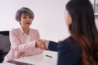 Deneyimli Asyalı profesyonel kadın ve genç yetişkin kadın ortaklık başarısını simgeleyen yeni bir iş teklifini mühürlemek için el sıkışıyor..