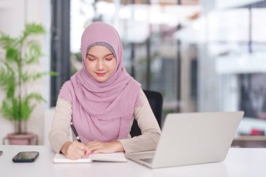 Odaklanmış Asyalı Müslüman kadın modern ofis masasındaki iş büyümesi için dizüstü bilgisayarla fikirleri özenle kaydediyor.