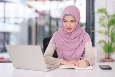 Ofis masasında çalışan tesettürlü yetişkin Müslüman kadın. O bir dizüstü bilgisayar kullanıyor ve bir deftere yazıyor. Bu verimliliği ve kendini işine adamış.