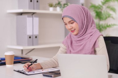 Odaklanmış Asyalı Müslüman girişimci, parlak çağdaş ofis ortamında bir dizüstü bilgisayar kullanarak ayrıntılı mali raporlar hazırlıyor