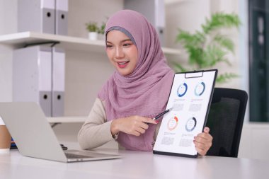 Kendine güvenen genç Müslüman profesyonel kadın, parlak bir ofis ortamındaki sanal sunum sırasında çizelgeleri bir panoda analiz ediyor..