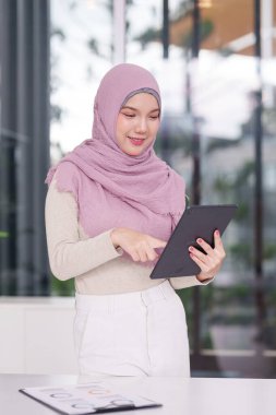 İş için dijital tabletle meşgul olan gülümseyen genç Asyalı Müslüman kadın. Modern ofis ortamında verileri analiz ediyor. Profesyonel başarılı gelecek odaklı.