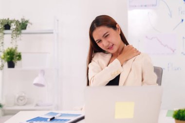 Asyalı iş kadını ofis masasında dizüstü bilgisayar kullanarak omuzlarında ve boynunda rahatsızlık hissediyor.