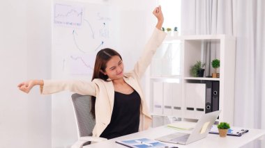 Asyalı profesyonel kadın temiz ofis masasında dinleniyor. Çalışma alanı iş başarısını ve kârını gösteren çizelgeler gösteriyor. Bu görüntü modern bir çalışma ortamında rahatlık ve verimliliği temsil eder..