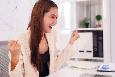 Asyalı yetişkin kadın modern ofis ortamında iş başarısından çok memnun. Kariyerinin başarısını kutluyor. Zafer kazanmış ve motive olmuş hissediyor.