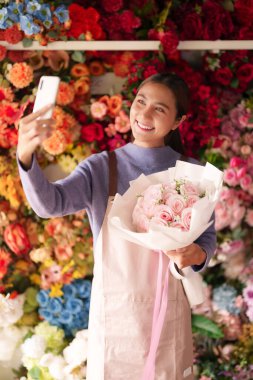 Mutlu Asyalı yetişkin çiçekçi elinde pembe gül buketiyle selfie çeker. Renkli çiçekçi atmosferinde dijital içerik yaratıcısı meşgul ediliyor.