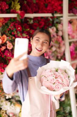Neşeli Asyalı çiçekçi, canlı çiçekçi dükkanında çiçekçilik sergileyen akıllı telefonuyla güzel pembe güller yakalıyor..