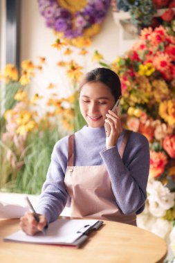 Genç Asyalı yetişkin kadın çiçekçi, telefon üzerinden müşteri siparişi alırken özenle notlar yazarak çiçekçi dükkanını verimli bir şekilde yönetiyor.