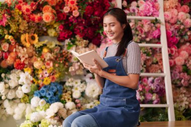 Asyalı yetişkin çiçekçi kadın canlı çiçekçi dükkanında çalışıyor defter okuyarak çiçek tasarımı düzenlemeleri yapıyor. Etrafı güzel çiçeklerle çevrili günlük işlerle uğraşıyor..