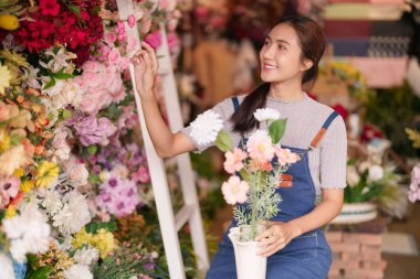 Asyalı yetişkin kadın aktif olarak yapay çiçekleri düzenliyor. Çiçek dükkanı sahibi gayretli bir şekilde canlı ürünler sergiliyor. Çiçek sergisinde neşeli bayan profesyonel çalışmalar.