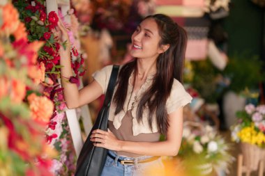 Genç Asyalı kadın renkli çiçekleri keşfederken gülümsüyor parlak bir çiçekçi dükkanında. İlham ve mutluluğu yeni çiçek aranjmanlarını seçerken buluyor..