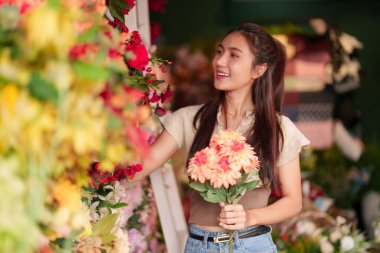 Yetişkin Asyalı bir kadın çiçek butiğinde canlı çiçekleri keşfeder. Özel bir düzenleme için çiçek seçerken elinde neşeli bir buket tutar.