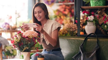 Gülümseyen Asyalı kadın, canlı bir çiçekçi dükkanında akıllı telefonuyla büyüleyici çiçek aranjmanlarını fotoğraflıyor.