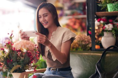 Gülümseyen Asyalı kadın telefonuyla güzel çiçekleri yakalıyor. Parlak, büyüleyici bir çiçekçinin içinde. Mutlu bir hava yaratıyor.