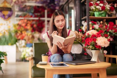 Asyalı kadın canlı bir çiçekçide çalışmaya odaklanmış güzel çiçekler ve bitkilerle çevrili gelişen işi için notlar alıyor.