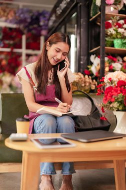 Asyalı yetişkin kadın profesyonel çiçekçi, çiçekçide çalışıyor müşteri arıyor iş planları yazıyor, günlük perakende operasyonlarını yönetiyor.