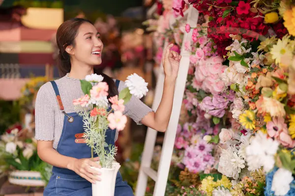 Neşeli Asyalı kadın çiçekçi yetenekli bir şekilde hareketli bir dükkanda canlı çiçekler düzenler müşteriler için eşsiz buketler yaratır.