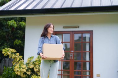 Mutlu Asyalı kadın hayallerindeki eve taşınıyor elinde bir karton kutu, modern bir evin önünde.