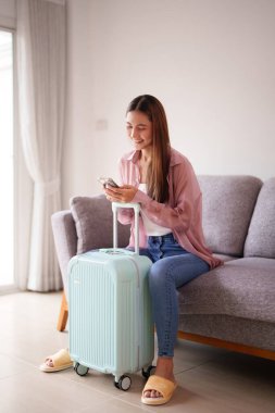 Mutlu Asyalı yetişkin kadın tatilini cep telefonuyla planlıyor. Açık yeşil bavulu heyecanlı bir yolculuk bekliyor..