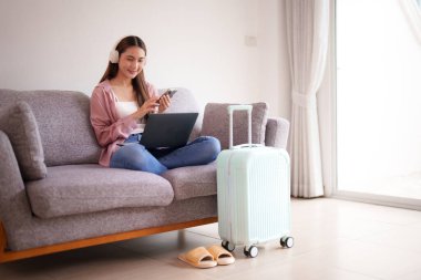 Rahat yetişkin Asyalı kadın uzaktan seyahat etmeyi planlıyor. Bilgisayarlı akıllı telefon ve kulaklık gibi modern teknolojiler kullanıyor..