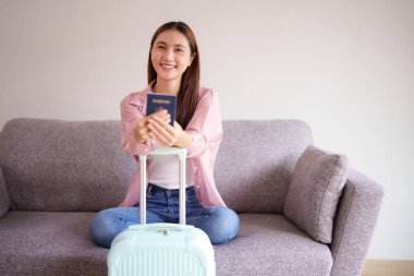 Gülümseyen Asyalı kadın pasaportunu heyecan verici bir yolculuğa hazırladı. Açık mavi bavul yakınlarda duruyor. Yaklaşan tatil tatili ev beklentisi.