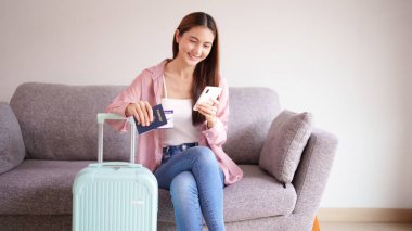Asyalı yetişkin kadın mutlu bir şekilde tatile hazırlanıyor. Elinde pasaportuyla seyahat detaylarını ve seyahat kartlarını kontrol ediyor. Heyecan verici bir yolculuğa hazır.