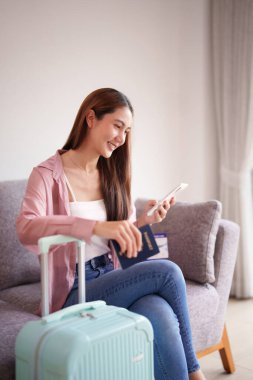 Gülümseyen Asyalı yetişkin kadın, çantasının yanında cep telefonu pasaportuyla seyahat etmeyi planlıyor.