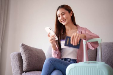 Gülümseyen Asyalı kadın seyahat için hazırlanıyor. Elinde pasaport ve telefon rezervasyonu ile rahat bir koltukta oturuyor.