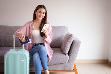 Neşeli Asyalı kadın uluslararası seyahat detaylarını tamamladı. Kalkmadan önce koltukta rahatça dinlenirken pasaportunu ve akıllı telefonunu tutuyor..