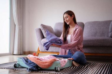 Yetişkin Asyalı kadın evinde giysi eşyasıyla seyahat çantasını neşeyle hazırlıyor. Yaklaşan bir tatil ya da macera için dikkatlice hazırlanıyor..