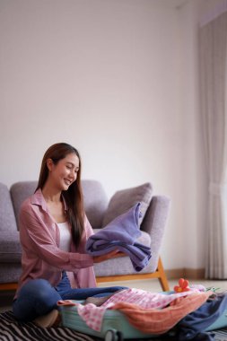 Mutlu Asyalı kadın rahat evindeki bavullarına giysilerini doldurmak için yolculuğa hazırlanıyor. Gülümsüyor. Seyahat planından hoşlanıyor..