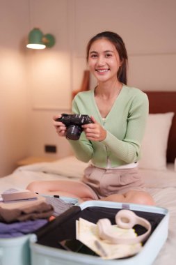 Neşeli Asyalı kadın, bavulunu kamera kulaklıkları ve yaklaşan tatil için kıyafet gibi seyahat gereksinimleriyle dolu heyecanlı bir maceraya hazırlanıyor.